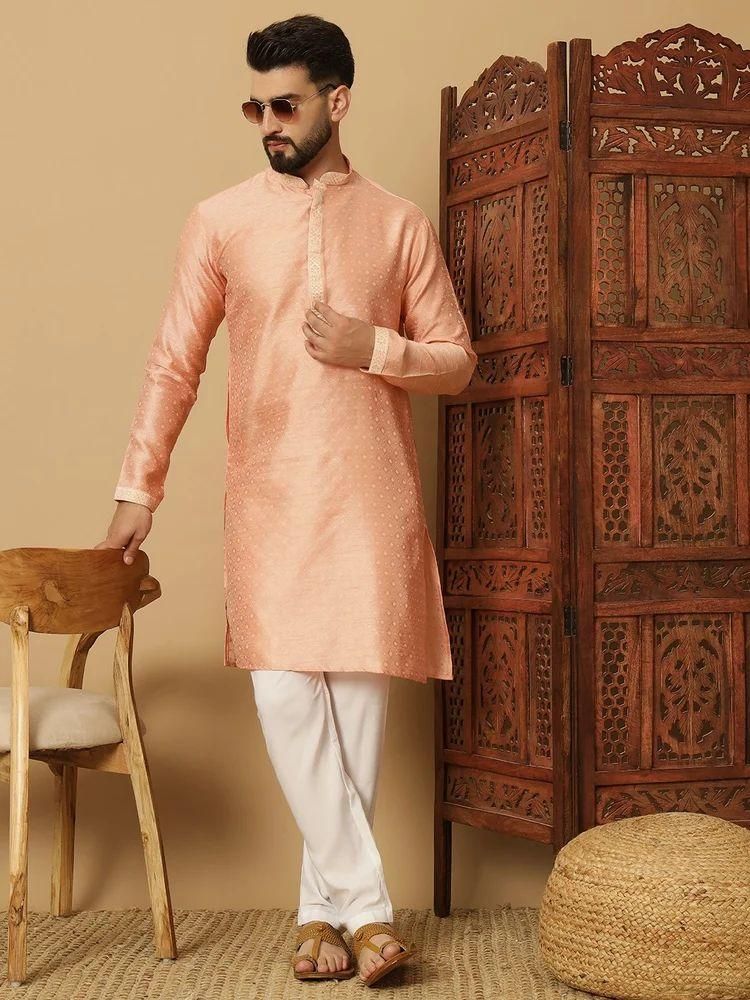 Man wearing a peach kurta with white pants standing next to a wooden chair and carved wooden screen.