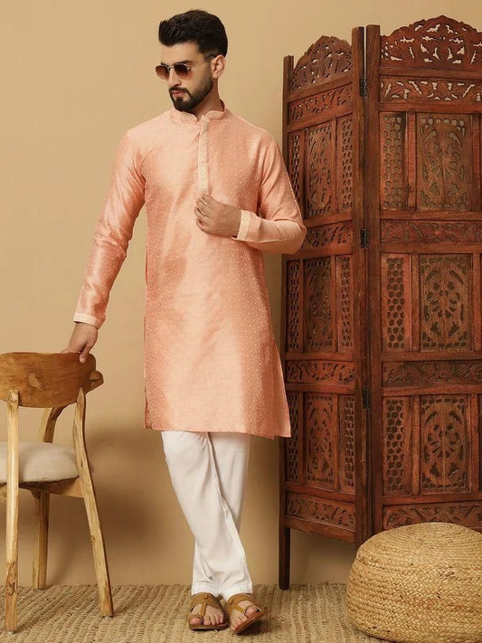 Man wearing a peach kurta with white pants standing next to a wooden chair and carved wooden screen.